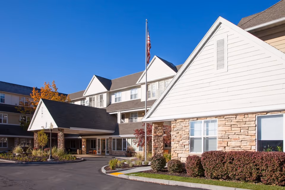 Front exterior of The Wellington senior living facility showing the entrance canopy, flagpole with an American flag, and landscaped driveway.
