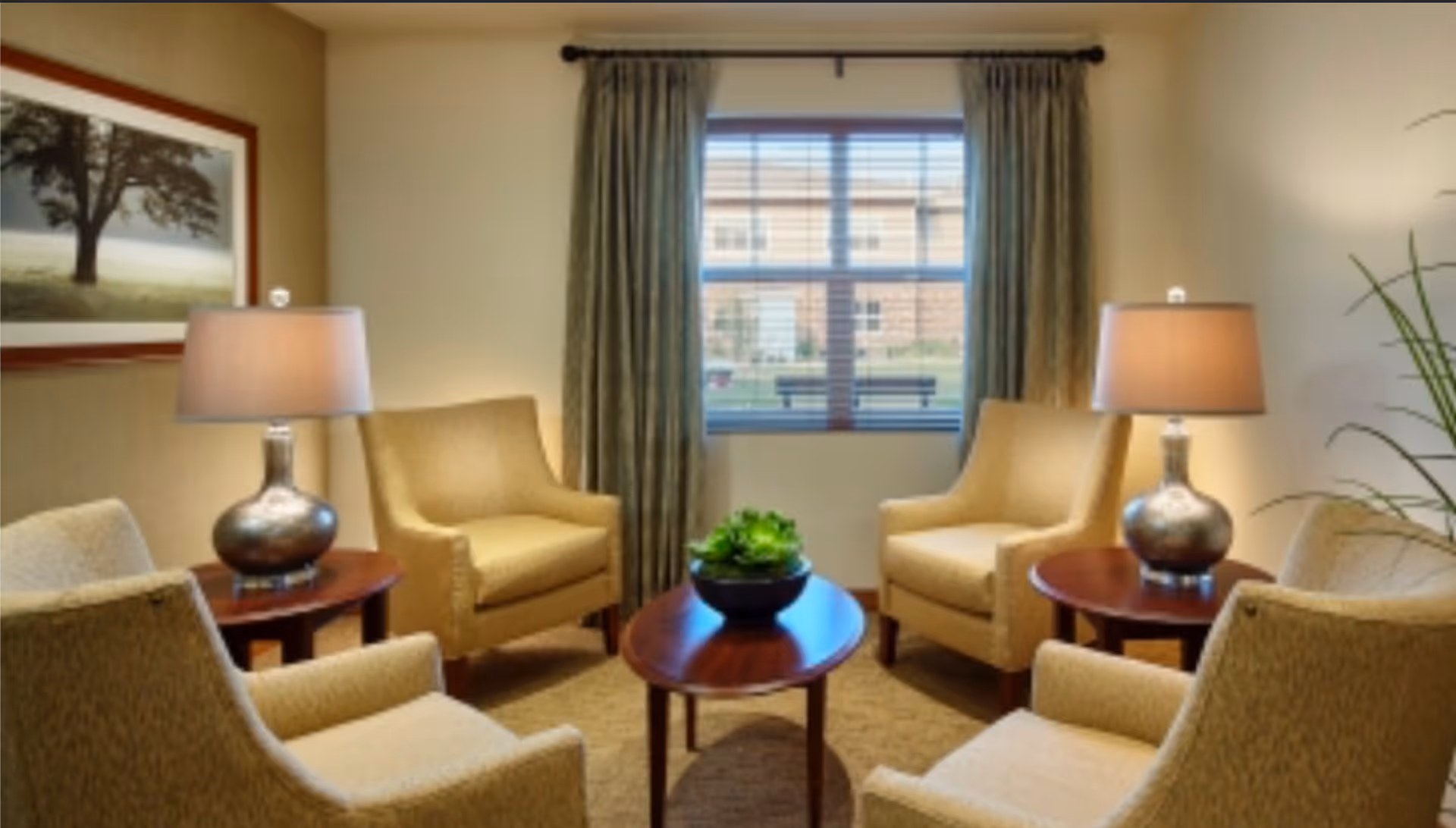 A cozy sitting area with four beige armchairs arranged around a wooden oval coffee table with a green plant centerpiece. Two side tables each hold a lamp with a metallic base and beige shade. A large window with green curtains lets in natural light, and a framed picture of a tree hangs on the wall.