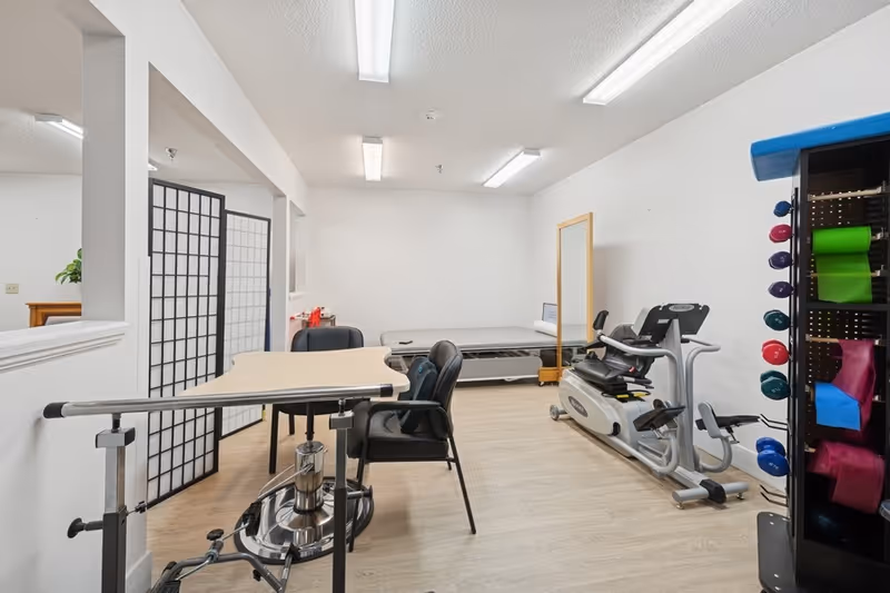 A therapy or rehabilitation room with exercise equipment including a recumbent bike, a therapy bed, a large mirror, and a rack holding colorful exercise bands and weights. There is also a table with chairs and a folding screen divider in the background. The room has white walls and bright overhead lighting.