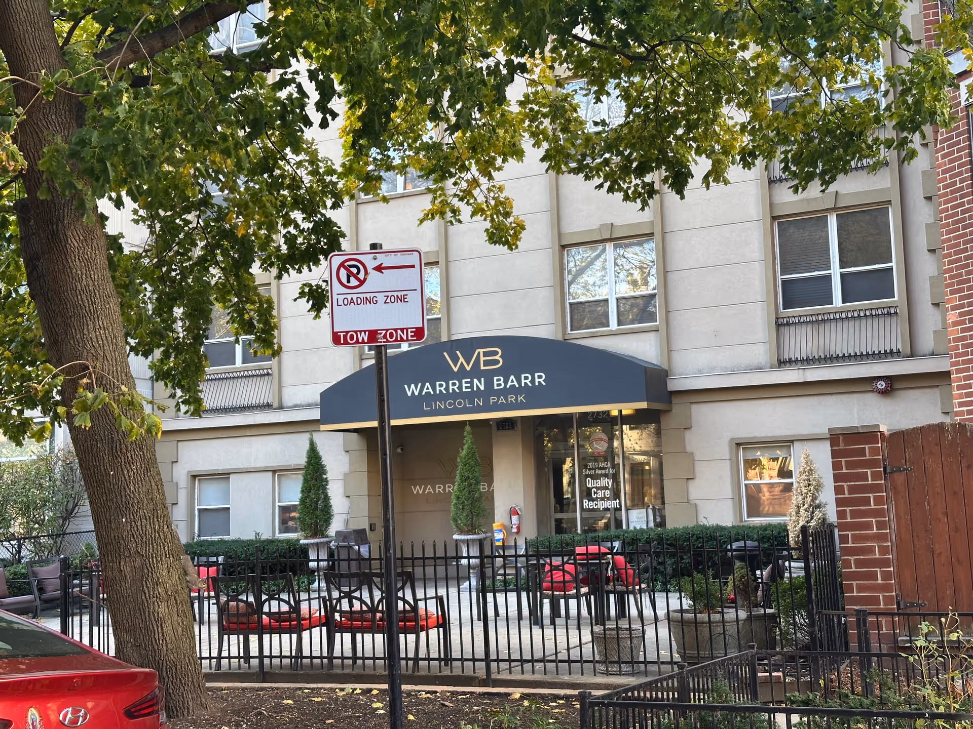 Front entrance of Warren Barr Lincoln Park with a black awning, fenced patio seating, trees, and a loading zone sign.