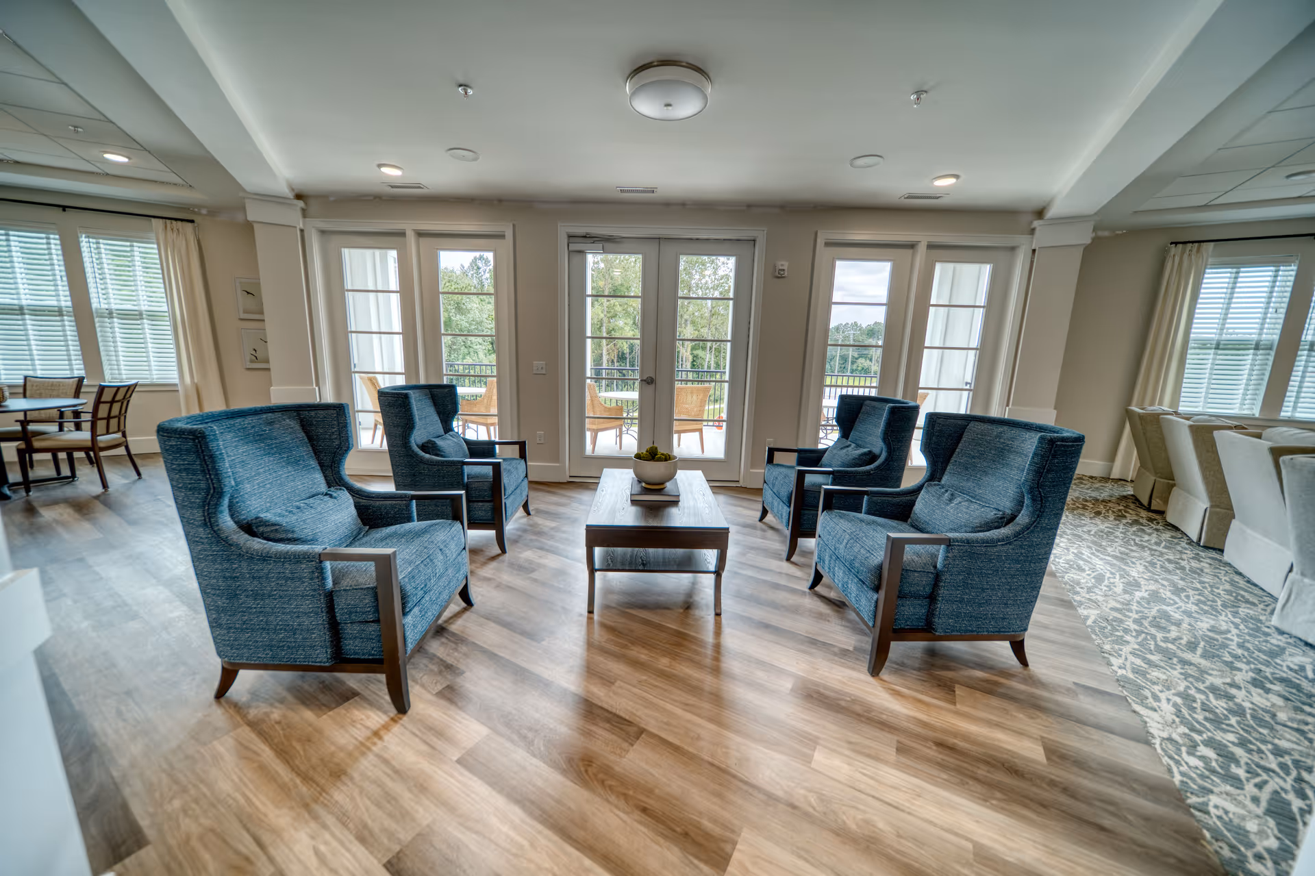 A bright and spacious living room area with four blue upholstered armchairs arranged around a wooden coffee table. Large windows and glass doors let in natural light and provide a view of an outdoor patio with chairs and greenery beyond. The room features wood flooring and light-colored walls with curtains on the windows.