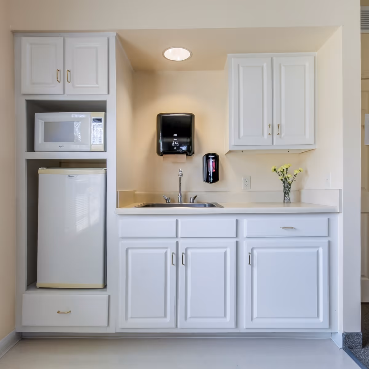 A small kitchenette area with white cabinets, a countertop with a sink, a microwave, and a mini refrigerator. There is a black paper towel dispenser and a black soap dispenser mounted on the wall above the sink. A small vase with yellow flowers is placed on the countertop.