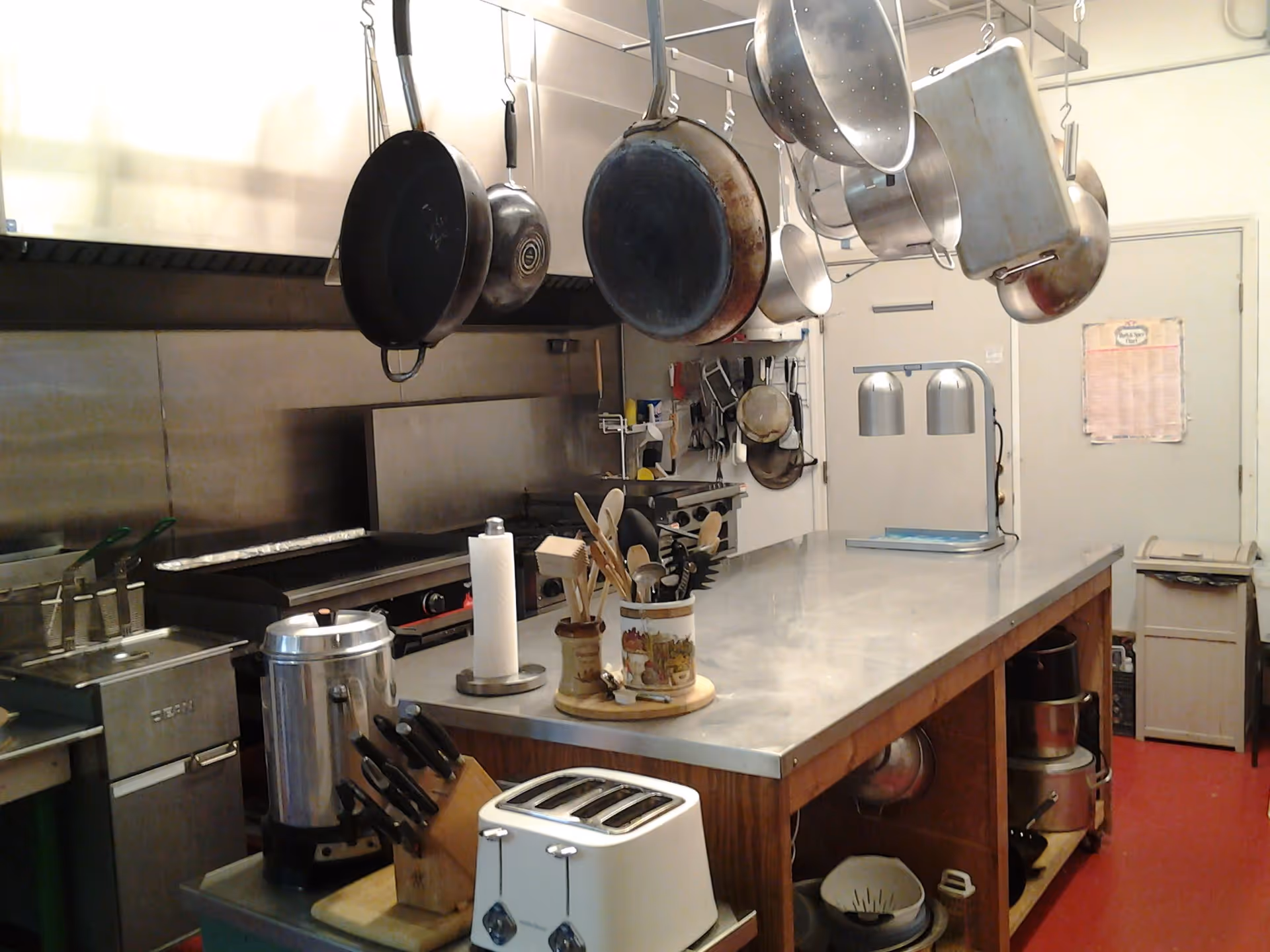 A commercial kitchen with hanging pans above a stainless-steel prep island stocked with utensils, a toaster, and other cooking equipment.