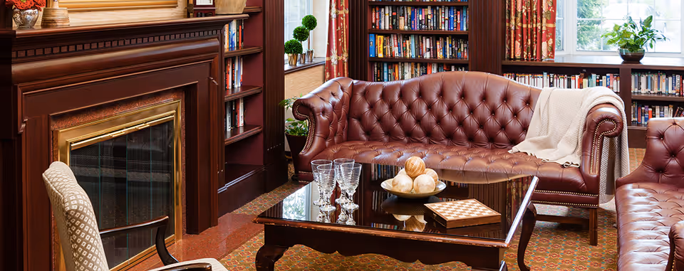 A cozy living room with a brown leather tufted sofa and matching armchair, a glass-top coffee table with decorative balls and a chessboard, a fireplace with a wooden mantel, bookshelves filled with books, patterned curtains, and a window with potted plants.