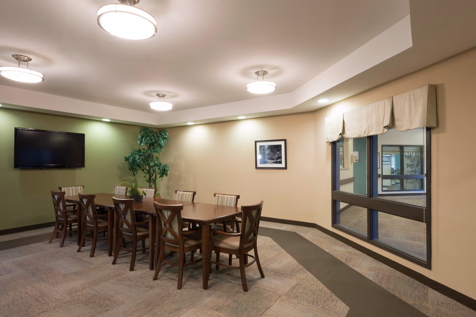 A well-lit room with a long wooden table surrounded by ten chairs. The walls are painted in beige and green tones, with a flat-screen TV mounted on the green wall. There is a large window with beige valances, a potted plant in the corner, and a framed black and white photograph on the wall.