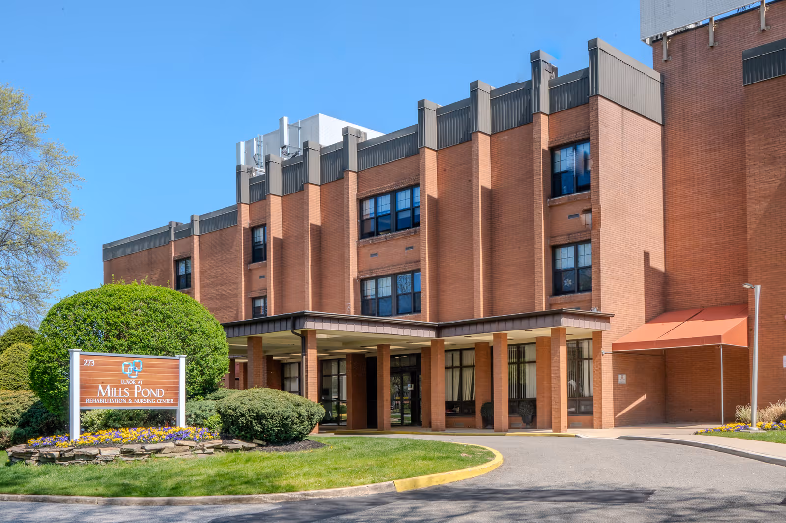 Exterior view of Luxor at Mills Pond Rehabilitation & Nursing Center, a multi-story brick building with large windows, an entrance canopy, and well-maintained landscaping including bushes and flowers under a clear blue sky.
