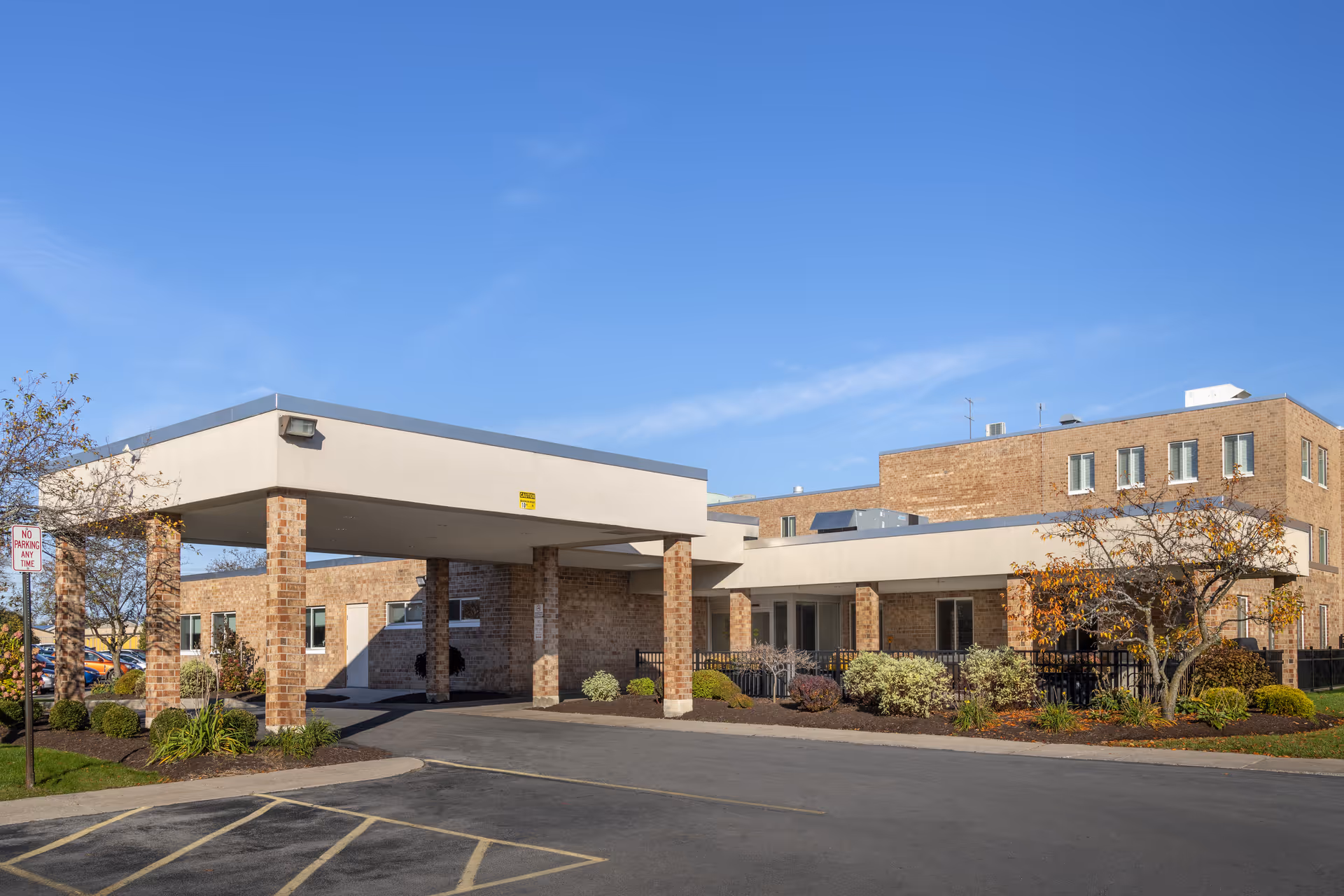 Exterior view of a brick senior living facility building with a covered entrance supported by brick pillars, surrounded by landscaped bushes and trees under a clear blue sky.