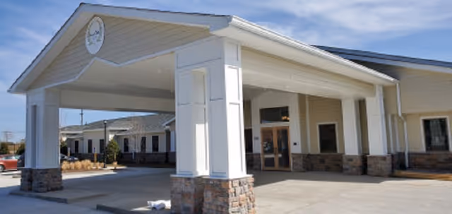 Exterior view of a single-story assisted living facility building with a covered entrance supported by white columns with stone bases, a clock mounted on the front gable, and clear blue sky above.
