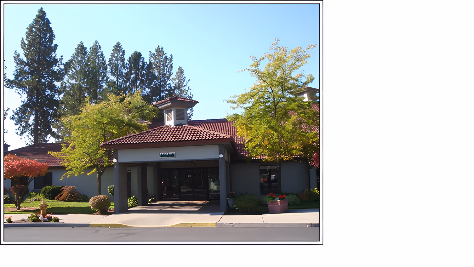 Front exterior view of a building with a red tiled roof, surrounded by green trees and shrubs under a clear blue sky.