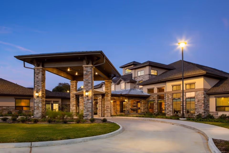 Exterior view of a senior living facility at dusk with a covered entrance supported by stone pillars, well-lit windows, and a curved driveway surrounded by landscaped greenery.