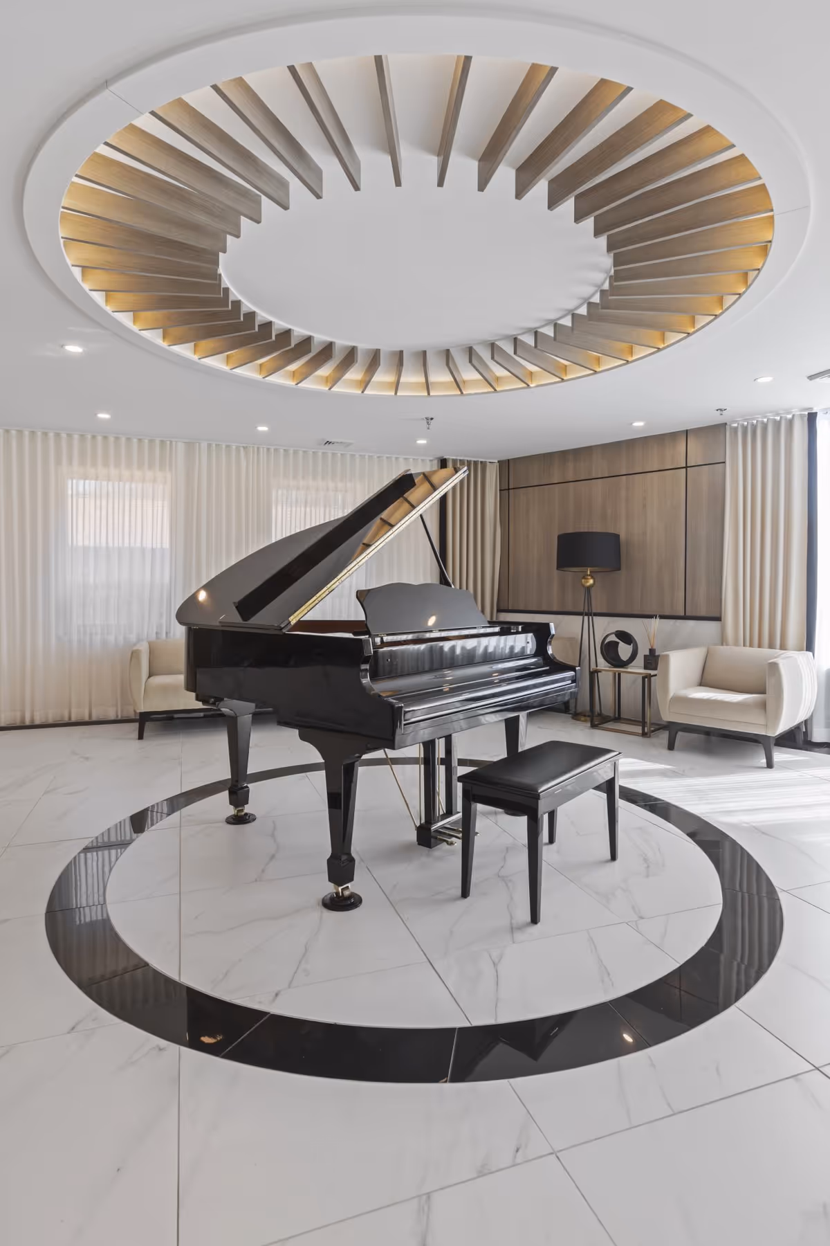 A modern interior room featuring a black grand piano with a matching bench placed on a circular patterned marble floor. The room has light-colored curtains, two beige armchairs, a side table with decorative items, a floor lamp, and a unique circular ceiling design with wooden slats and recessed lighting.