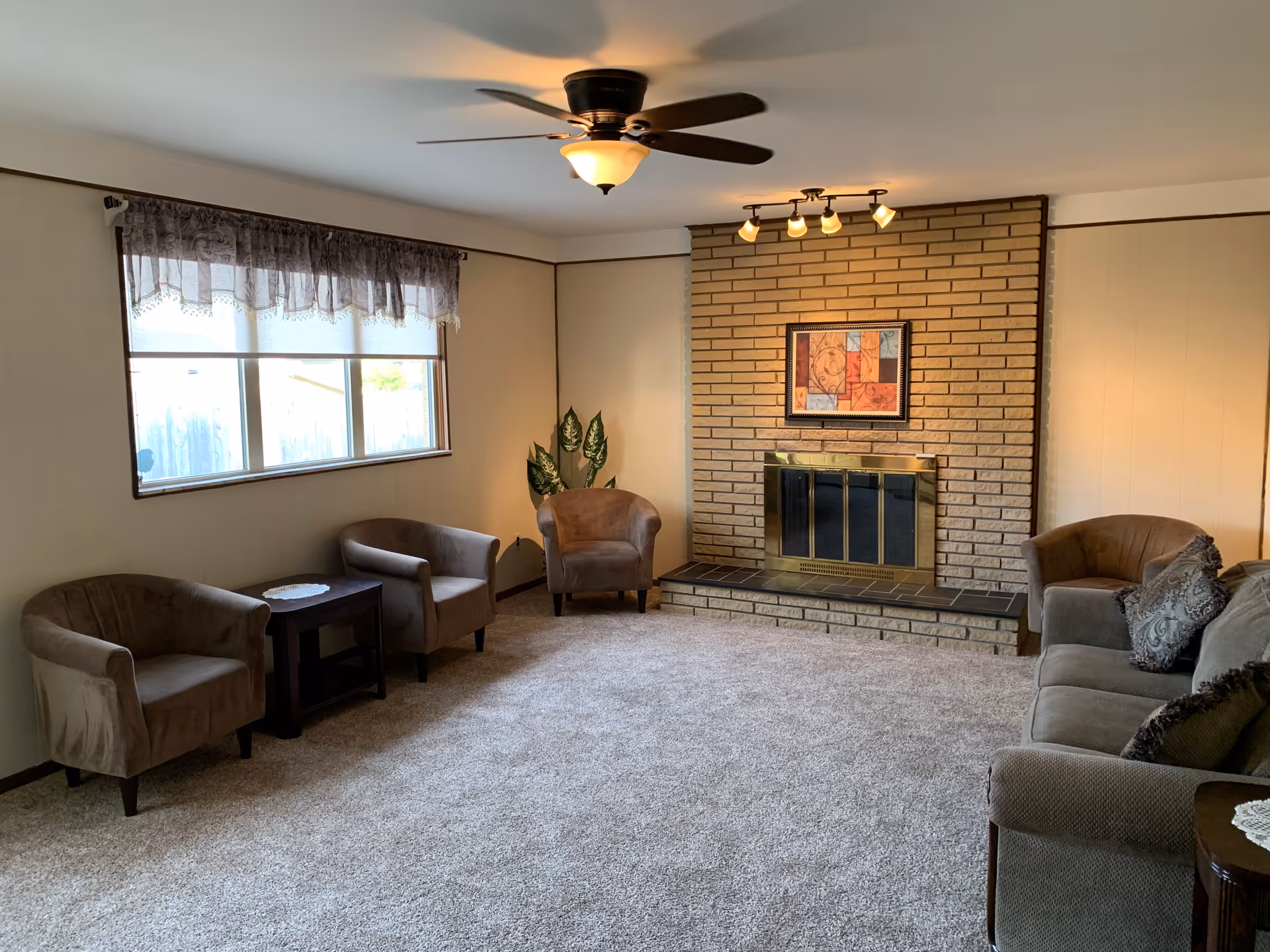 A cozy living room with beige carpet, a brick fireplace with a brass screen, and a framed artwork above it. The room has a ceiling fan with a light fixture, a window with a lace valance, three brown upholstered armchairs, a brown sofa with decorative pillows, a small dark wooden side table, and a green potted plant in the corner.