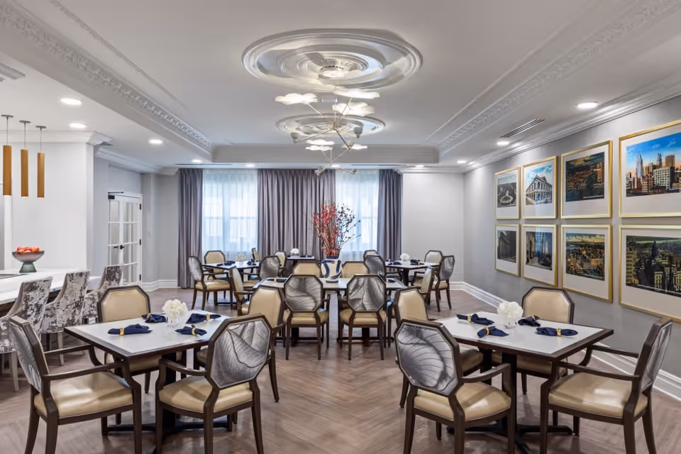 A bright and elegant dining room with multiple tables and chairs arranged neatly. The tables are set with navy blue napkins and white floral centerpieces. The room features a decorative ceiling with modern light fixtures, large windows with sheer curtains, and framed pictures on one wall. There is also a bar area with high chairs on the left side.