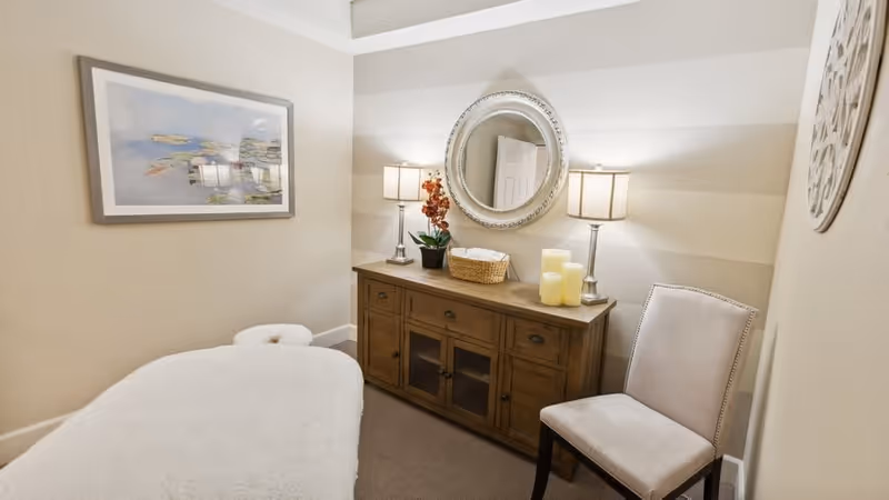 A cozy treatment room with a massage table covered in a white towel, a wooden cabinet with two lamps, a basket, candles, and a potted plant. A round mirror hangs on the striped wall above the cabinet, and a beige upholstered chair is placed beside it. A framed picture is on the adjacent wall.
