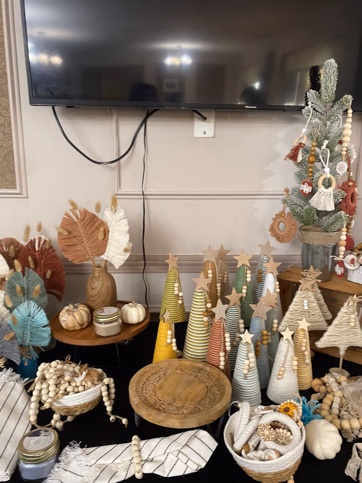 A decorative display featuring small handcrafted Christmas trees made of wrapped yarn in various colors with wooden star toppers and bead garlands, alongside small pumpkins, woven baskets, candles, and dried floral arrangements, set on a black tablecloth beneath a mounted flat-screen TV.