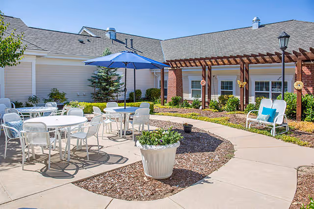 Outdoor patio area at Brookdale Highlands Ranch with white metal tables and chairs, a blue umbrella, a white planter with flowers, a curved concrete walkway, and a building with brick and siding in the background under a clear blue sky.
