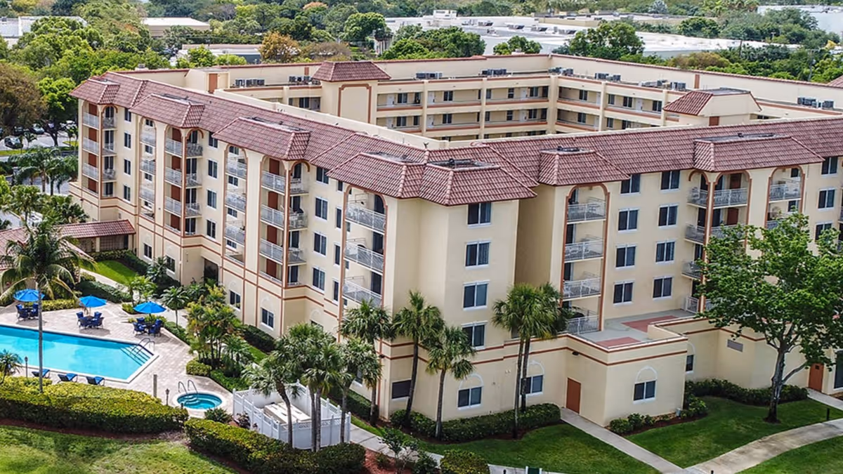 Aerial view of a multi-story senior living facility with beige walls and red-tiled roofs, surrounded by palm trees and greenery. The outdoor area features a swimming pool with blue umbrellas and lounge chairs, as well as a hot tub. The building has multiple balconies and is situated in a suburban area with other buildings and trees in the background.