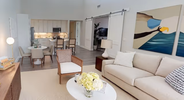 A bright and modern living area featuring a beige sofa with pillows, a wooden armchair with white cushion, and a white oval coffee table with a vase of flowers. Behind the living area is a dining space with a round table and four chairs, and a kitchen with light wood cabinets, stainless steel appliances, and a breakfast bar with two stools. The room has light walls, wood flooring in the kitchen and dining area, and carpet in the living area. A large abstract painting hangs on the wall above the sofa.