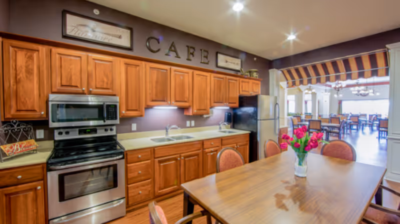 A kitchen area with wooden cabinets, a stainless steel oven and microwave, a double sink, and a refrigerator. A wooden table with chairs surrounds it, and a vase with red flowers is placed on the table. In the background, there is a dining area with multiple tables and chairs, and a striped awning above the entrance to the dining space. The word 'CAFE' is displayed on the wall above the cabinets.