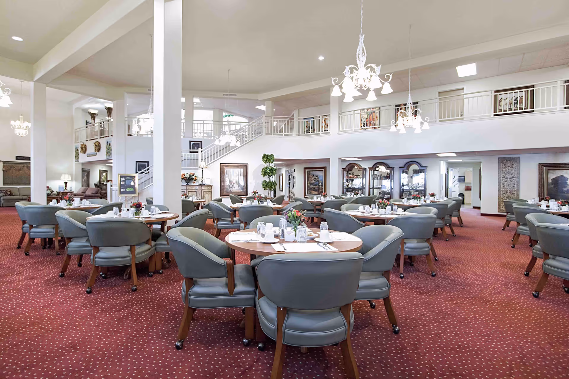 A spacious dining room with round tables set for meals, each table surrounded by cushioned chairs on wheels. The room features red carpet with a dotted pattern, white walls, and multiple chandeliers hanging from the ceiling. There is a staircase leading to an upper level with railings, and decorative plants and framed artwork adorn the space.