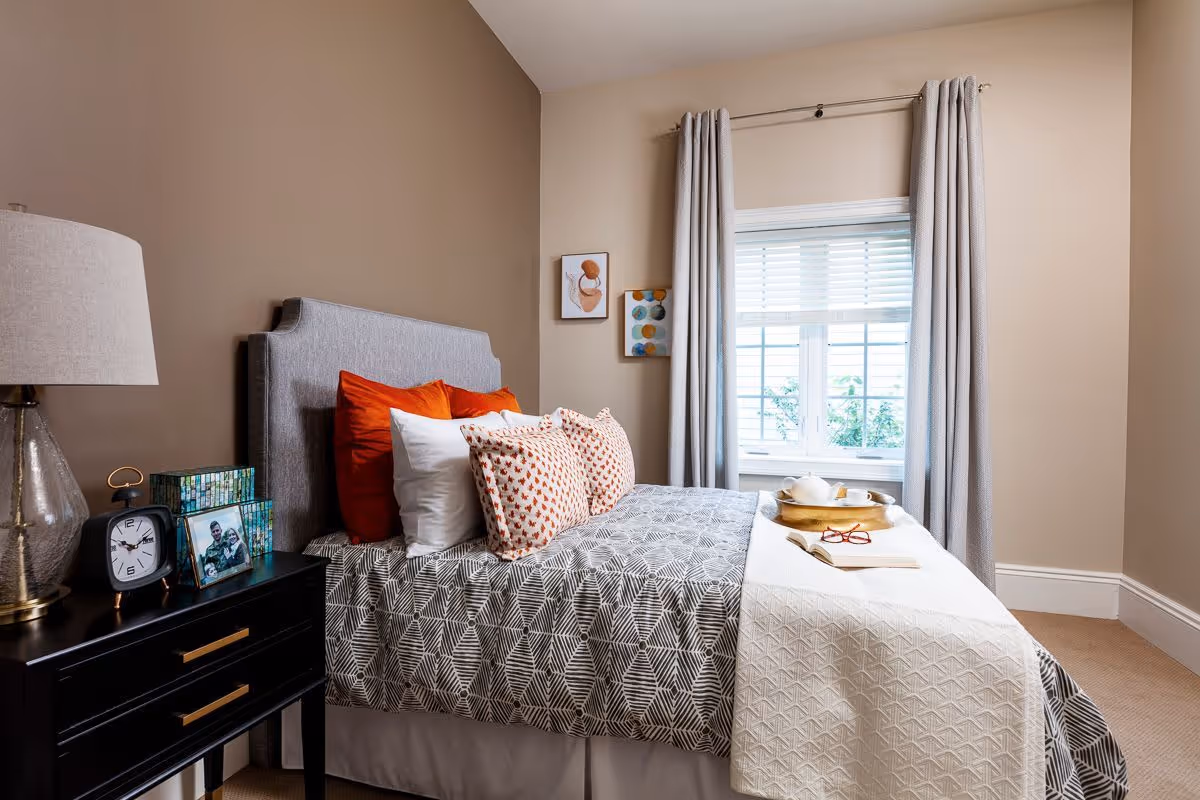 A cozy bedroom with a bed featuring a gray patterned bedspread, white and orange pillows, and a white throw blanket. A black nightstand with a lamp, clock, and framed photo is beside the bed. The room has beige walls, a window with gray curtains, and two small framed artworks on the wall.