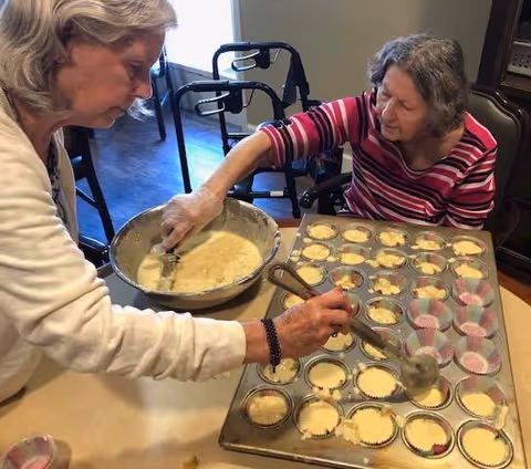 Two elderly women are preparing cupcakes together in a kitchen or dining area. One woman is scooping batter from a large bowl into cupcake liners in a muffin tray, while the other woman watches and assists. A walker is visible in the background.