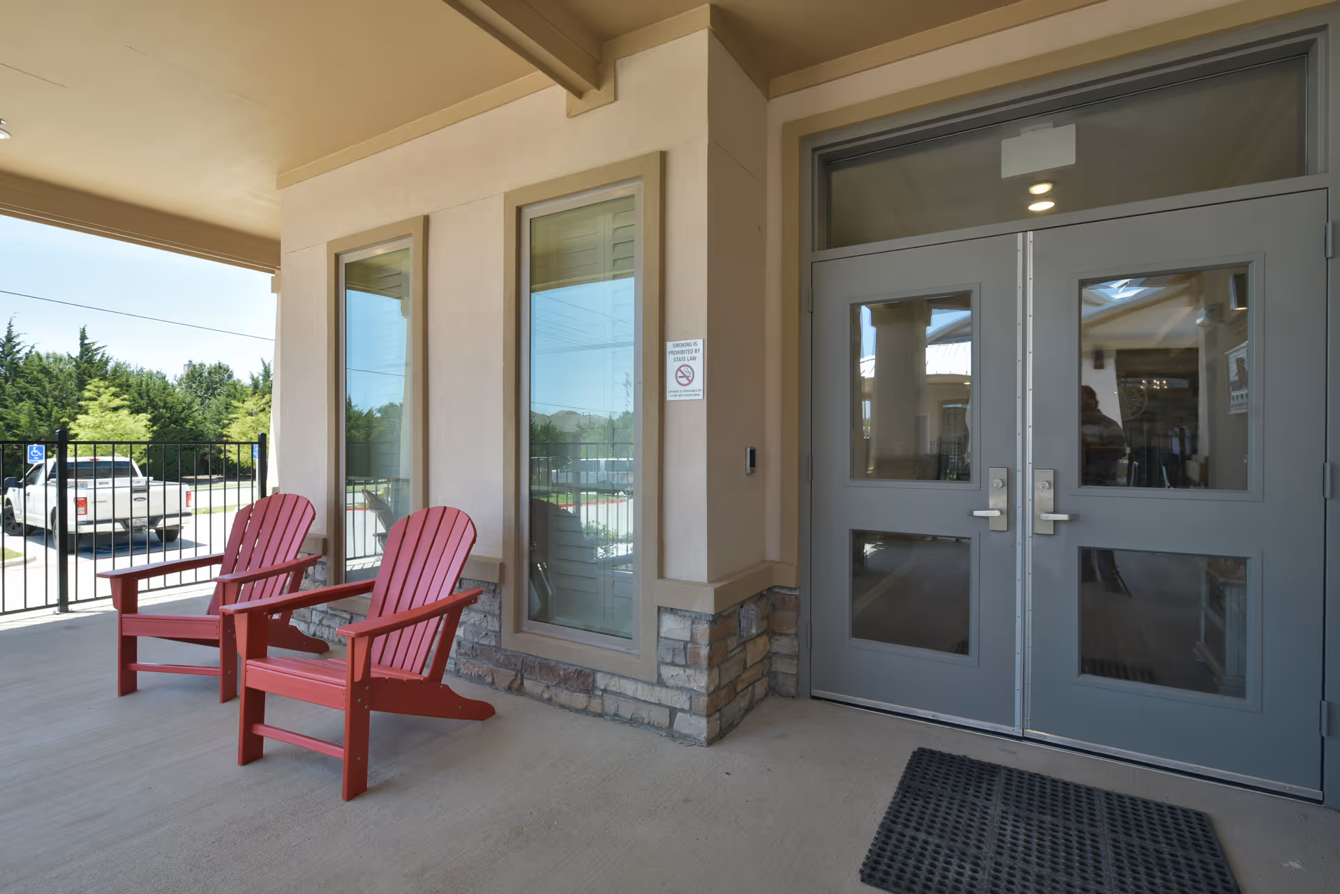 Covered outdoor entrance area of a facility with two red wooden chairs, two tall windows, a double gray door with glass panels, and a black rubber mat on the floor. A white pickup truck is visible in the background outside the fenced area.