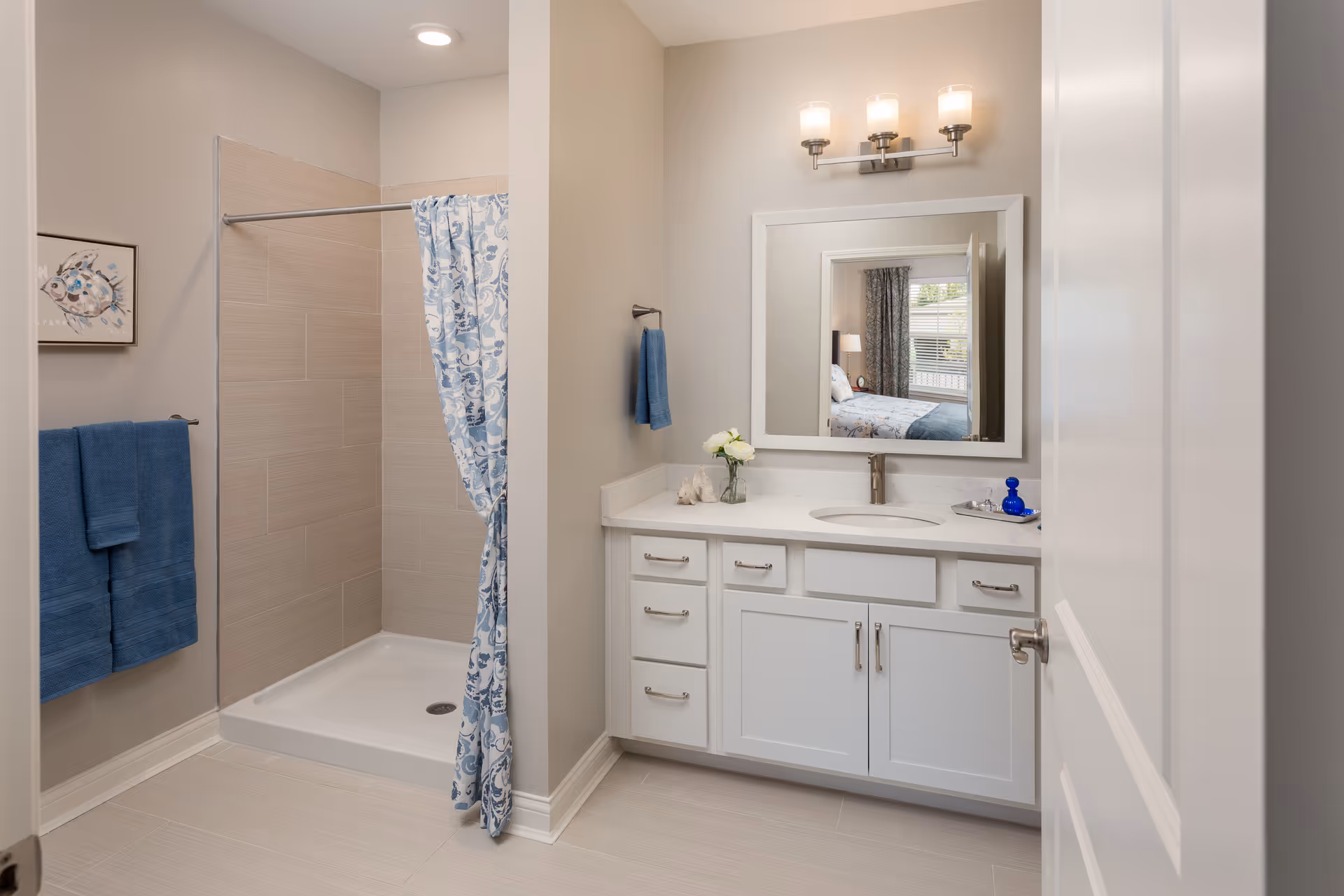A clean and modern bathroom with a walk-in shower featuring beige tiles and a blue and white patterned shower curtain. There is a white vanity with a sink, a large mirror above it, and a three-light fixture. Blue towels hang on the wall, and a small vase with white flowers is on the countertop. The reflection in the mirror shows a bedroom with a window and curtains.