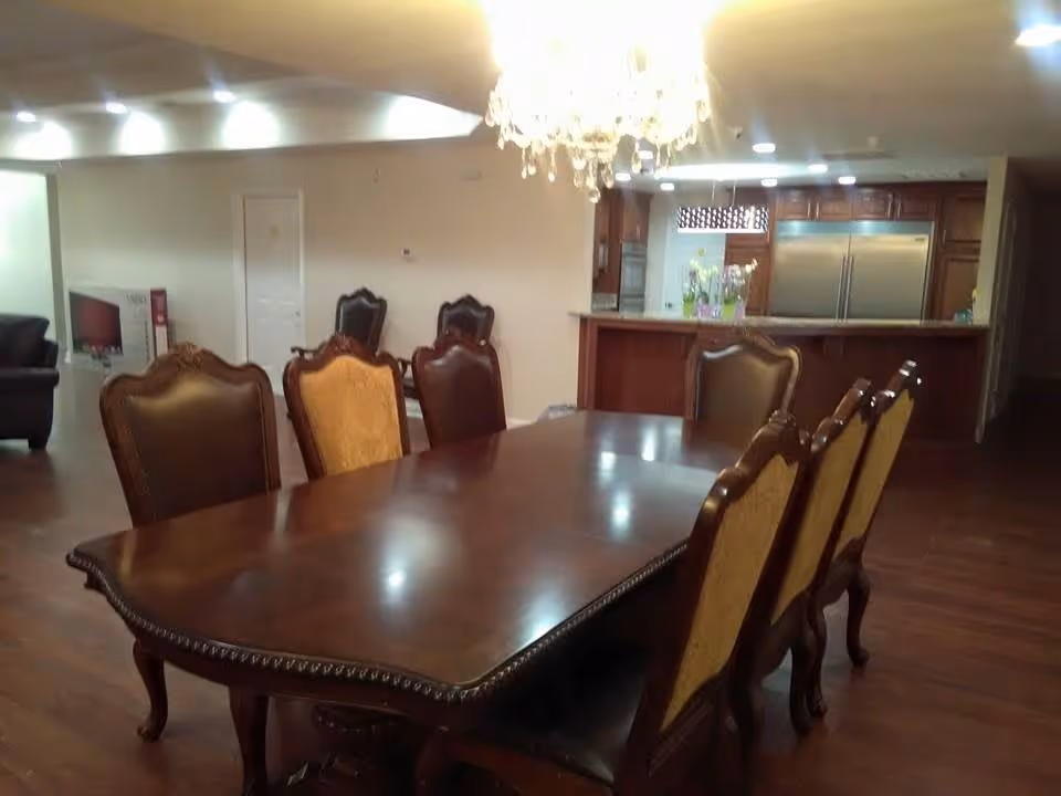 Interior view of a dining area with a large wooden dining table surrounded by eight ornate chairs, a chandelier hanging above, and a kitchen area with stainless steel refrigerator and wooden cabinets in the background.