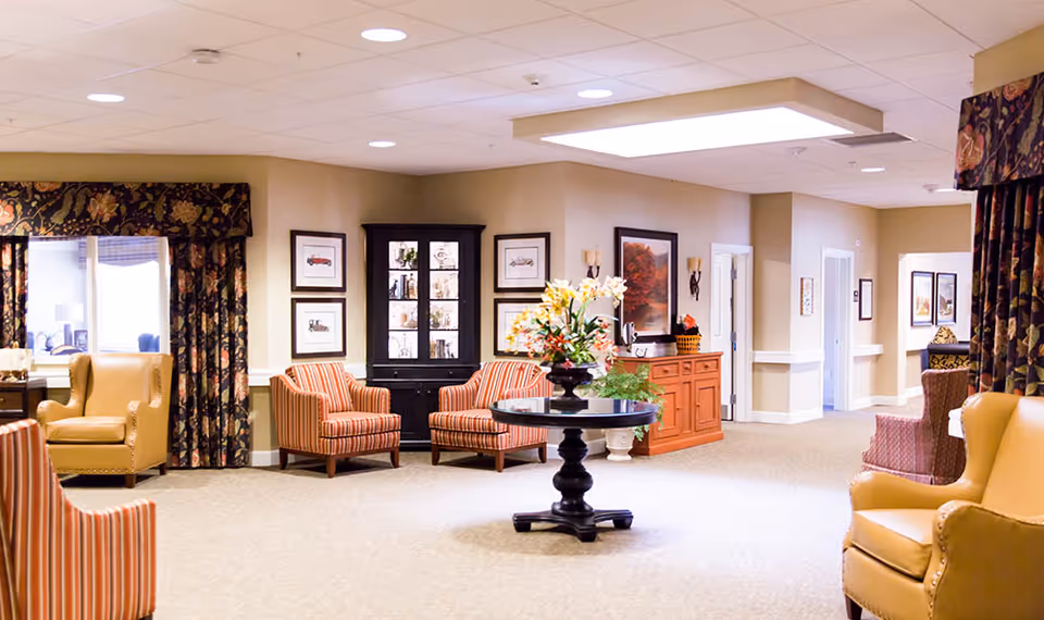 A spacious and well-lit common area in a senior living facility with beige walls and carpeted floors. The room features several comfortable armchairs and striped upholstered chairs arranged around a central round table with a floral arrangement. There are framed pictures on the walls, a black display cabinet, and a red sideboard with decorative items. Large windows with floral curtains allow natural light to enter the room.