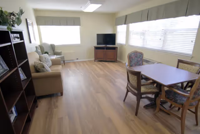 Bright communal living room with sofas and armchairs, a TV on a stand, and a table with chairs by large windows.