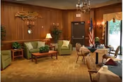 Wood-paneled community lounge with sofas, armchairs, small dining tables, a chandelier, and a U.S. flag by an exit door.