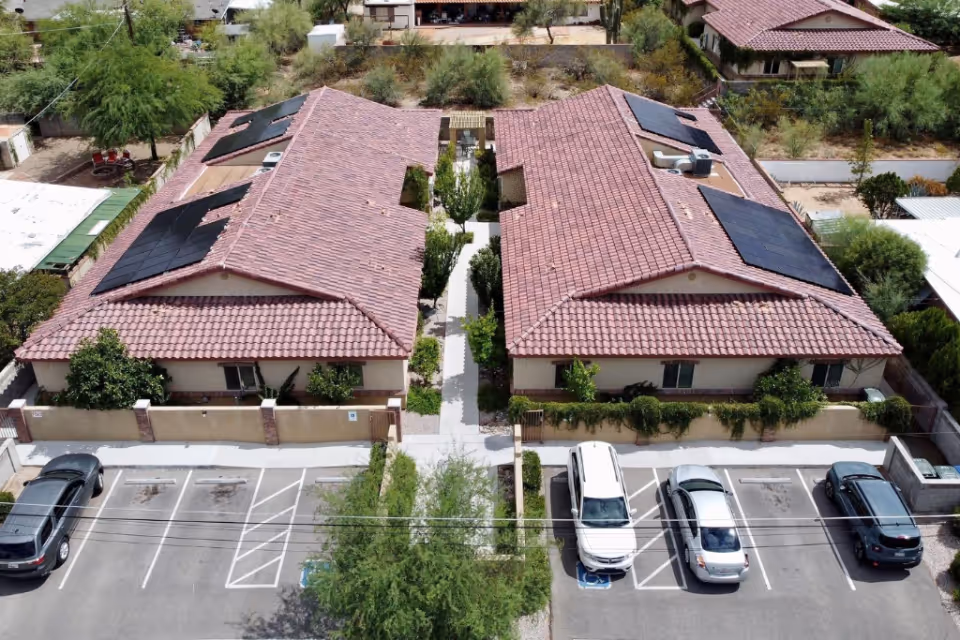 Aerial view of a residential care facility with two single-story buildings featuring red tiled roofs and solar panels. The buildings are separated by a walkway with greenery, and there are several parked cars in front of the facility. Surrounding the property are trees and other residential structures.