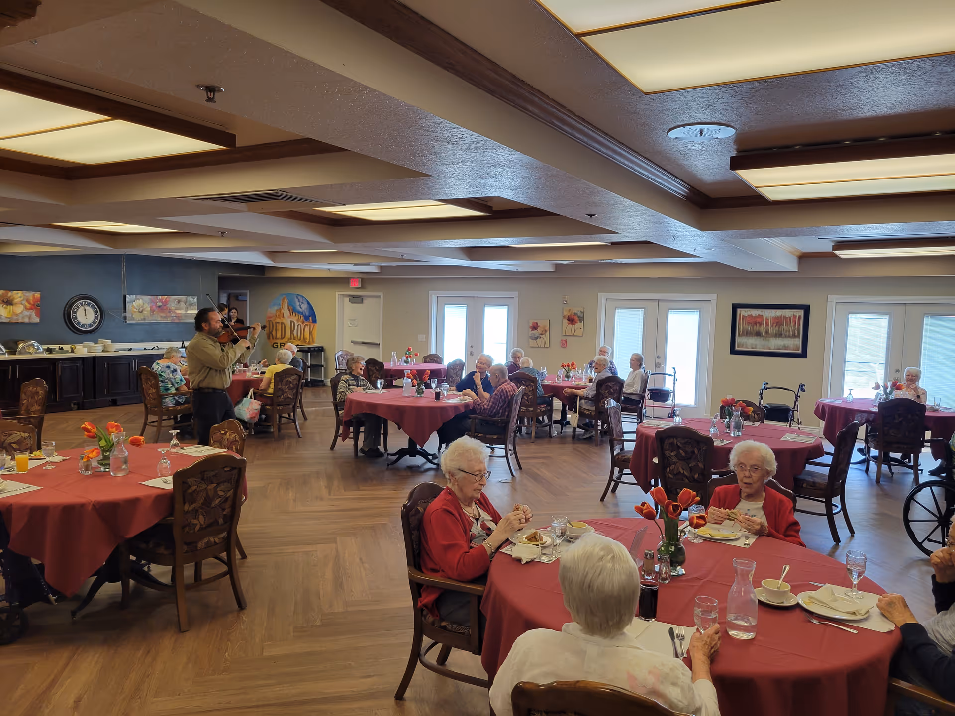 A dining room in an assisted living community with elderly residents seated at round tables covered with red tablecloths. The room has wooden flooring, ceiling lights, and wall decorations. A man is playing a violin near the back of the room, providing live music for the residents.