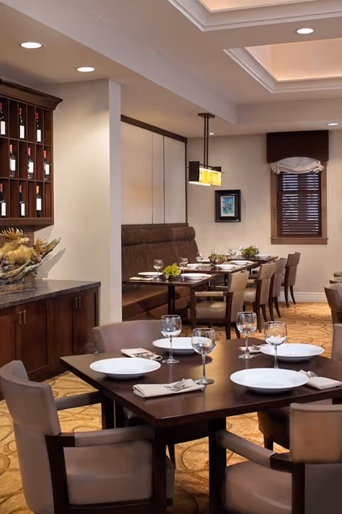 A well-lit dining room with neatly arranged tables set with white plates, wine glasses, and napkins. The room features comfortable upholstered chairs, a built-in wine rack on the left wall, and a window with wooden blinds. The ceiling has recessed lighting and a modern hanging light fixture above the tables.