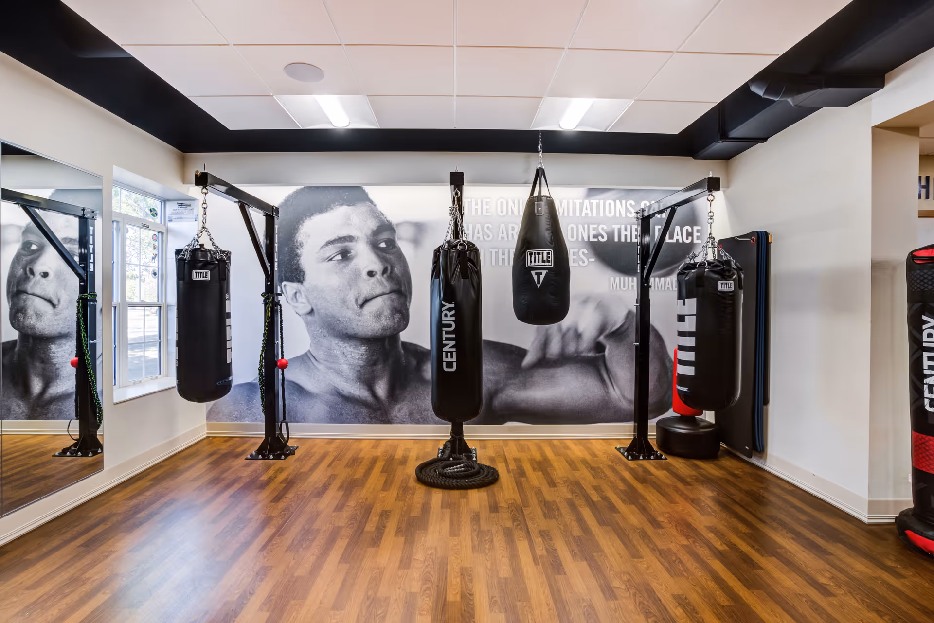 Interior view of a fitness room with wooden flooring, featuring four hanging punching bags and a large black and white mural of a boxer on the back wall. There is a window on the left side and a large mirror on the left wall reflecting part of the room.