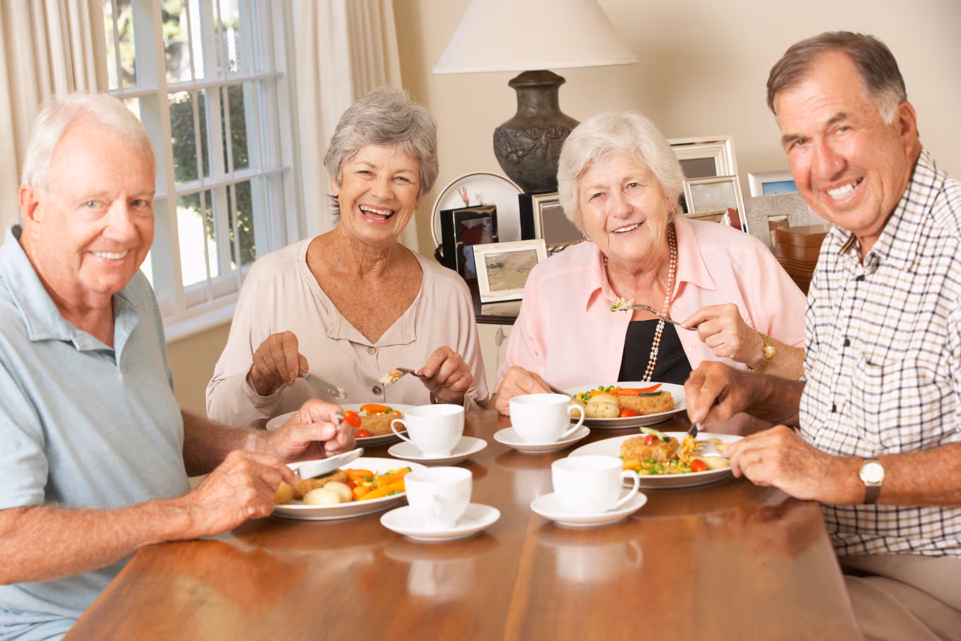 Four elderly people sitting around a dining table smiling while eating a meal.