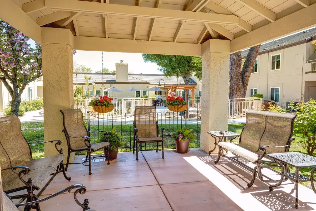 Covered outdoor seating area with metal chairs and benches, potted plants, and hanging flower baskets. In the background, there is a garden with trees, a fence, and a building with windows and umbrellas over tables.
