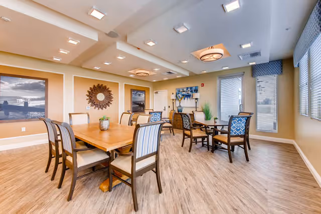 A bright and spacious dining room with wooden tables and cushioned chairs arranged neatly. The room features large windows with blinds allowing natural light to fill the space. The walls are decorated with framed artwork and a decorative mirror. The ceiling has recessed lighting and modern light fixtures.