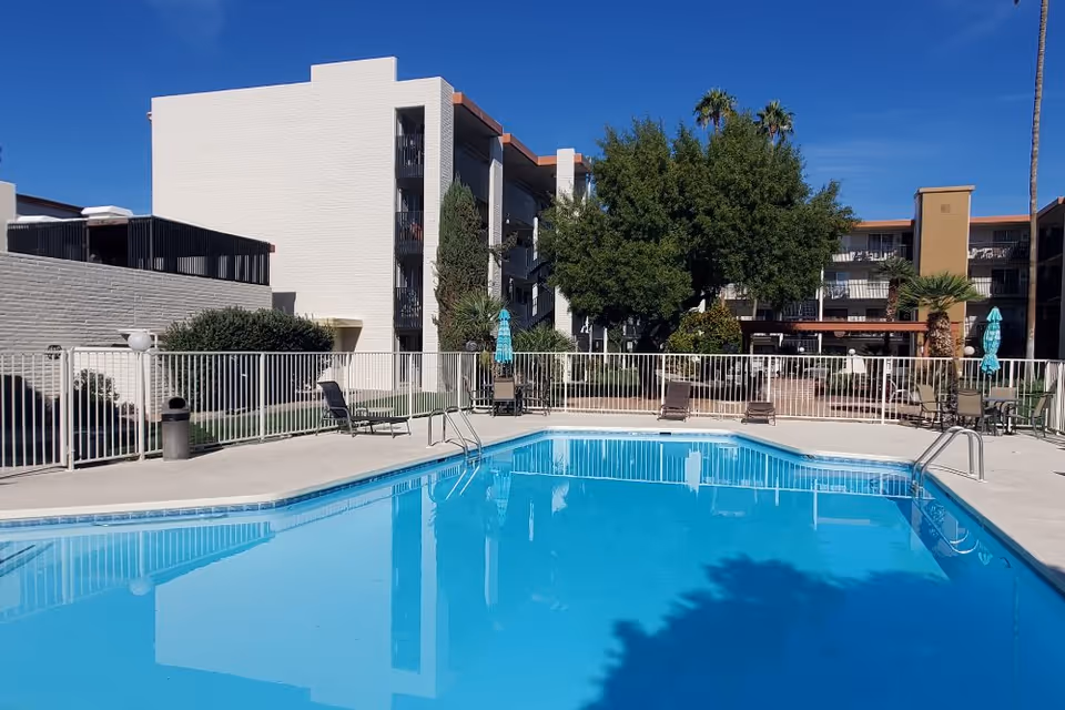 Outdoor swimming pool area at Fellowship Square Tucson with clear blue water, surrounded by a white safety fence. Several lounge chairs and tables with umbrellas are placed around the pool. In the background, there are multi-story residential buildings, trees, and a clear blue sky.