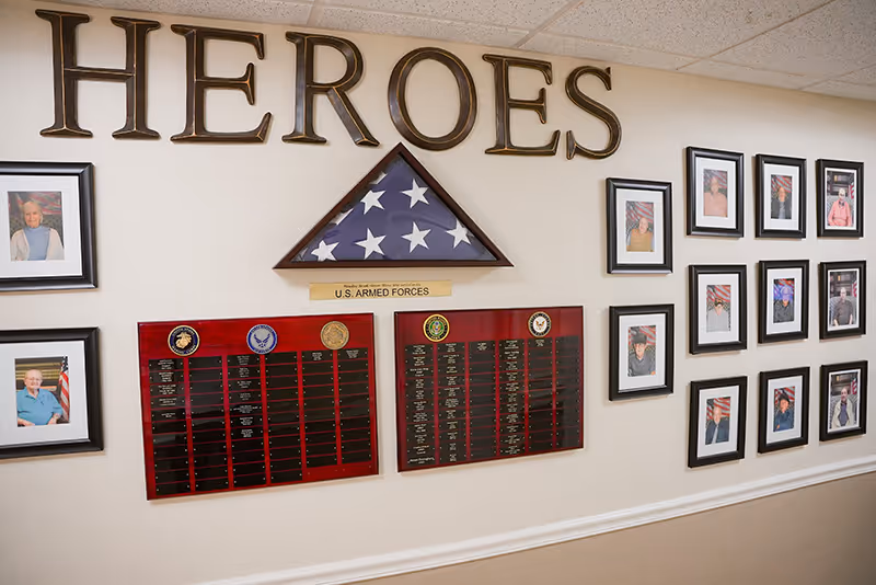A wall display honoring U.S. Armed Forces veterans with the word 'HEROES' in large letters above a folded American flag in a triangular case. Below the flag are two plaques with military branch emblems and names listed. Surrounding the plaques are framed photographs of veterans, each with an American flag background.