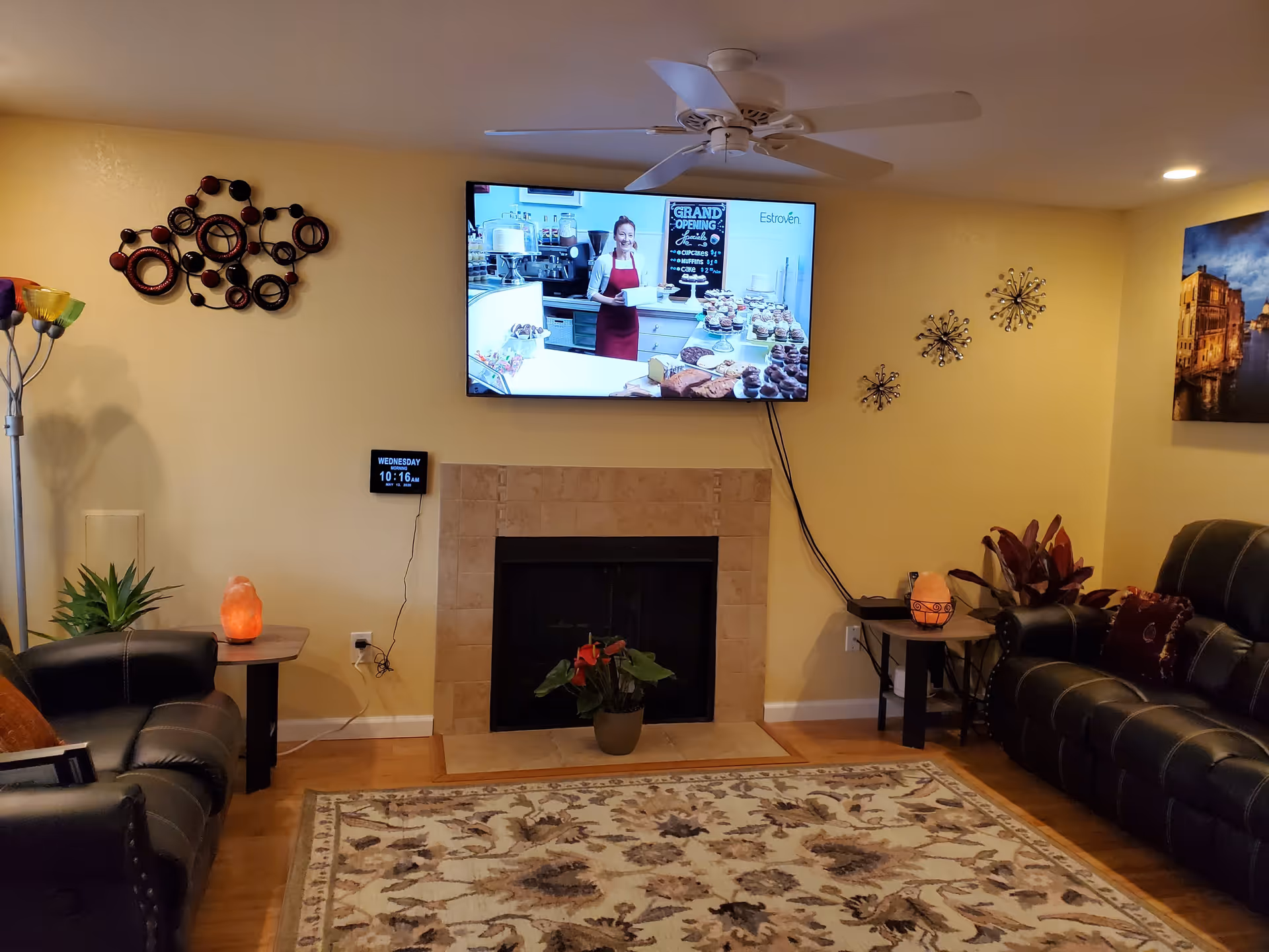 A cozy living room with yellow walls, a mounted flat-screen TV above a tiled fireplace, two black leather sofas on either side, side tables with salt lamps and plants, decorative wall art, a ceiling fan, and a patterned area rug on a wooden floor.
