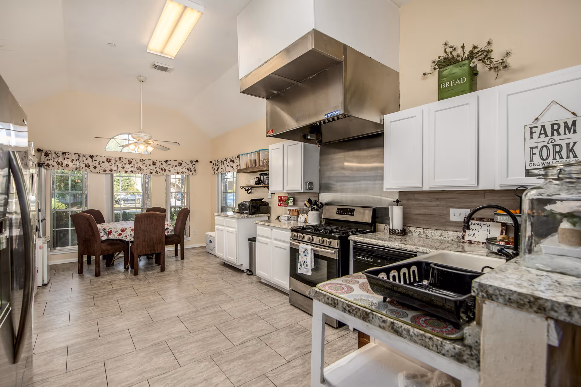 A bright kitchen and dining area with white cabinets, stainless steel appliances including a stove and refrigerator, a granite countertop, and a dining table with four brown upholstered chairs near large windows with floral valances.