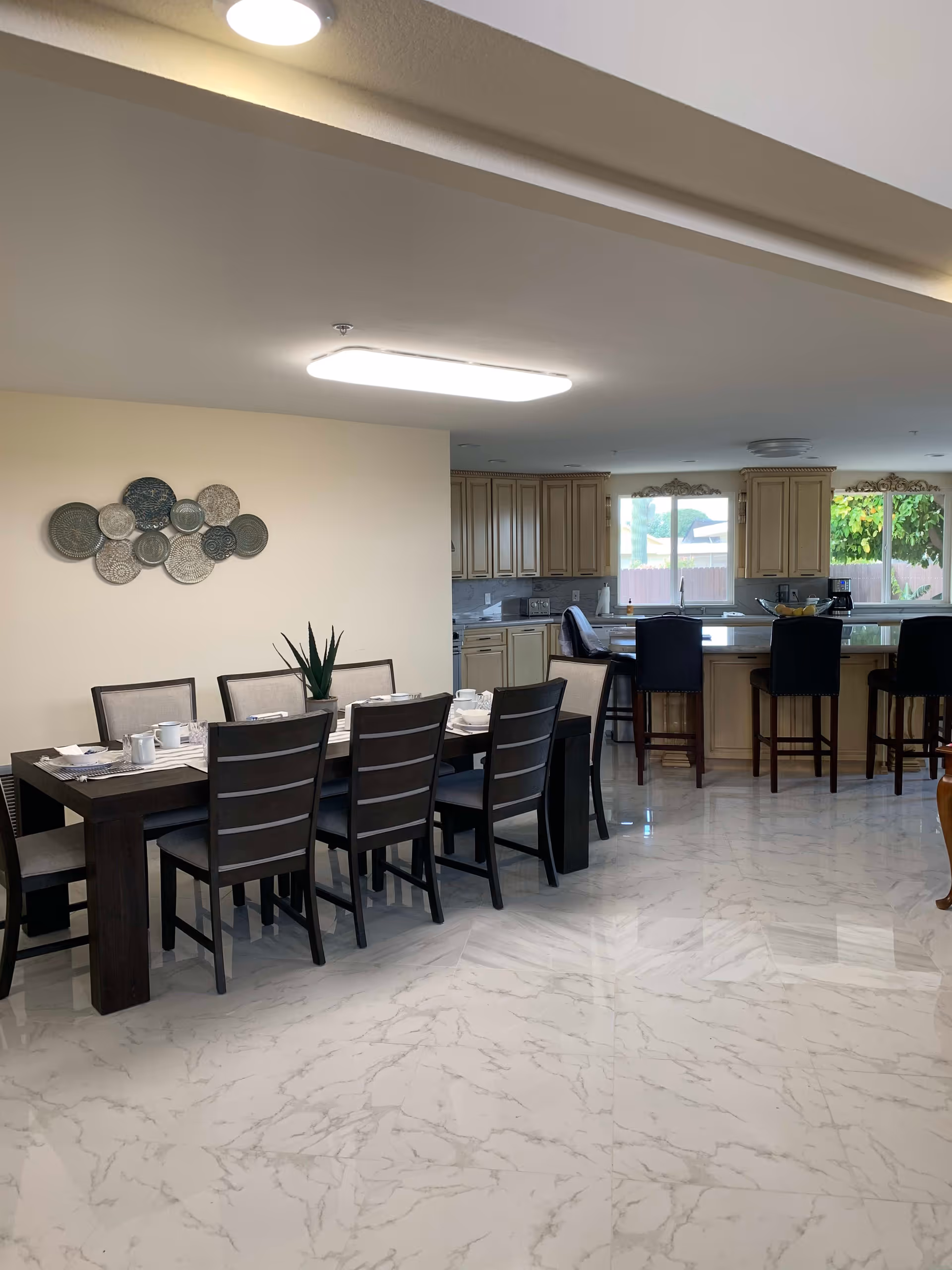 A spacious dining area with a long dark wooden dining table set for eight people with white dishes and cups. Behind the dining table is a kitchen with light-colored cabinets, a large island with four black bar stools, and two windows showing greenery outside. The floor is covered with white marble tiles, and there is decorative wall art above the dining table.