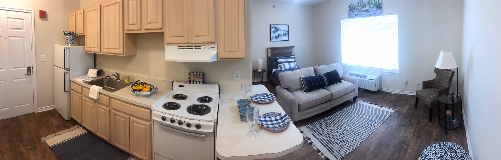 Open studio-style apartment interior with a small kitchen (stove, sink, refrigerator) and an adjacent living area featuring a sofa, chair, and window.