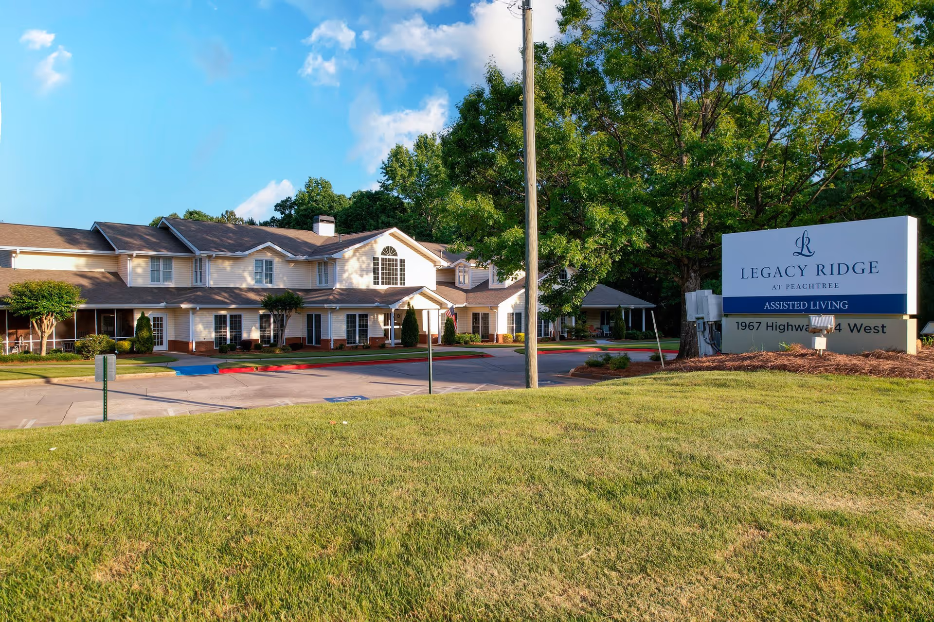 Exterior view of a two-story assisted living facility with beige siding and a brown roof, surrounded by green trees and a well-maintained lawn. A large sign in the foreground reads 'Legacy Ridge at Peachtree Assisted Living 1967 Highway 74 West'.