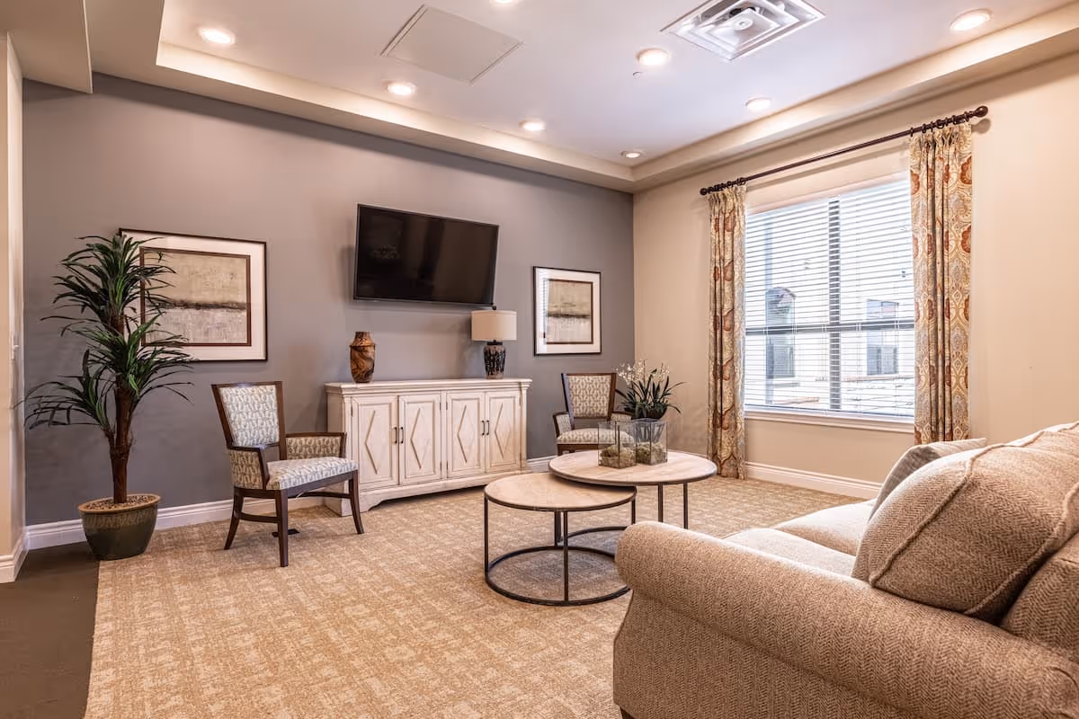 A cozy living room with a beige sofa, two patterned chairs, a white cabinet with decorative items, a wall-mounted flat-screen TV, a large window with patterned curtains, a potted plant, and two round nested coffee tables on a carpeted floor.