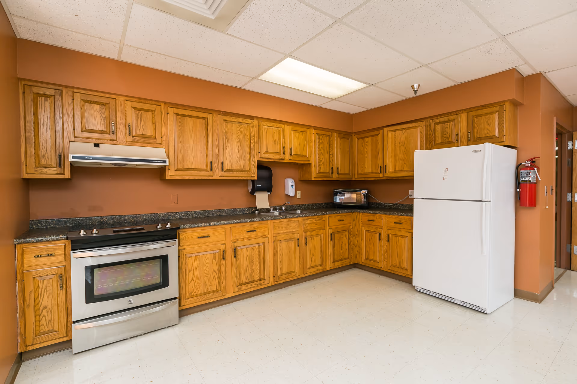 A spacious institutional kitchen with oak cabinets, granite countertops, a stainless stove and a white refrigerator.