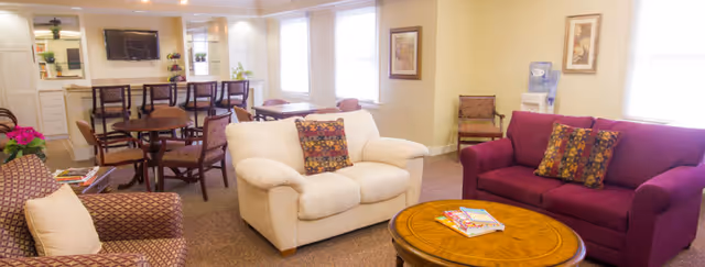 A cozy senior living facility common area with comfortable seating including a white loveseat, a purple sofa, and a patterned armchair. There is a round wooden coffee table with magazines on it in the center. In the background, there are several wooden chairs and tables, a wall-mounted TV, and large windows letting in natural light.