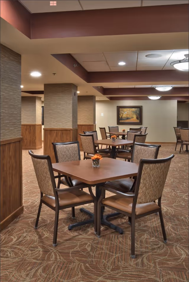 Interior view of a dining area with multiple square wooden tables and cushioned chairs arranged neatly. Each table has a small flower arrangement in a glass vase. The room features patterned carpet, beige walls with wood paneling, and ceiling lights. A framed painting of a tree-lined path hangs on the far wall.